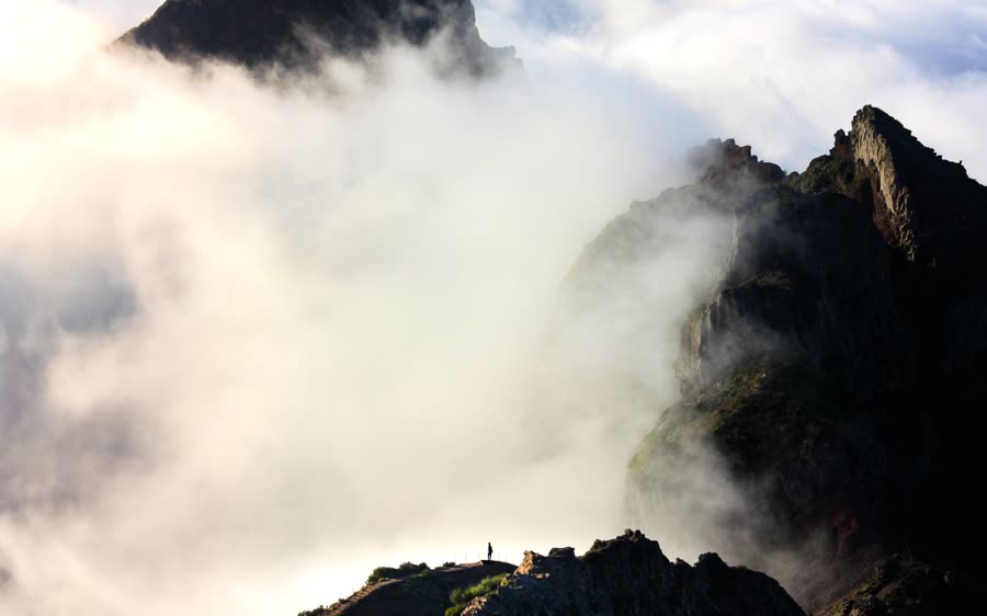 A lone hiker on a ridge, small against mountains rising out of the cloud