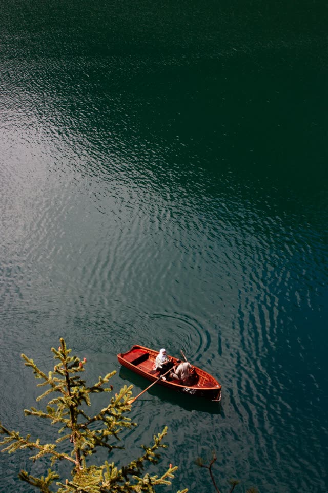 A red rowboat seen from above on clear green water, a pine branch in the corner