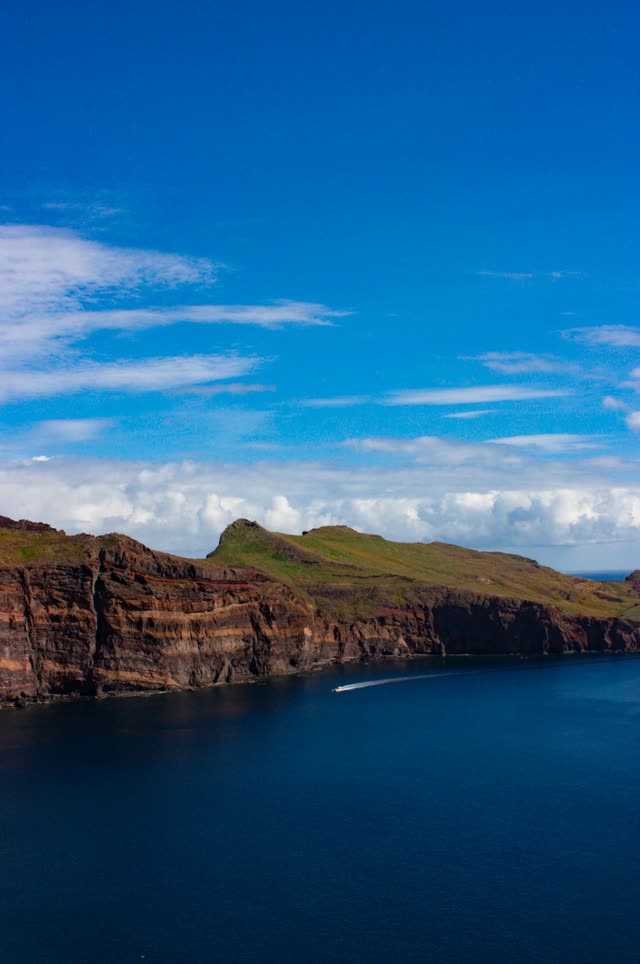 Layered red cliffs under a blue sky, a boat's wake cutting across the dark sea