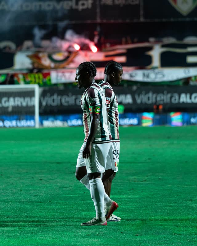 Two players walk past each other at night, a flare burning in the stand behind them