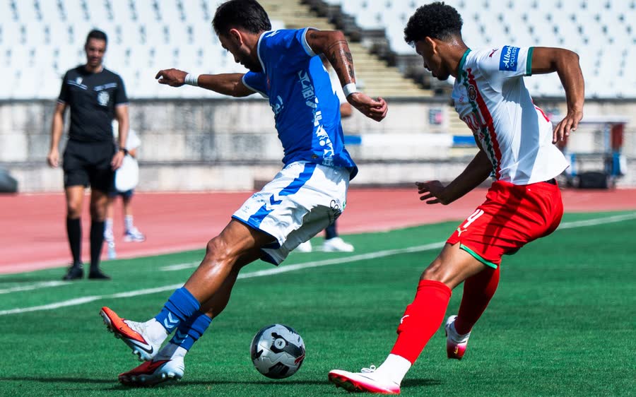 A player in blue shields the ball from a defender in white and red, a referee watching