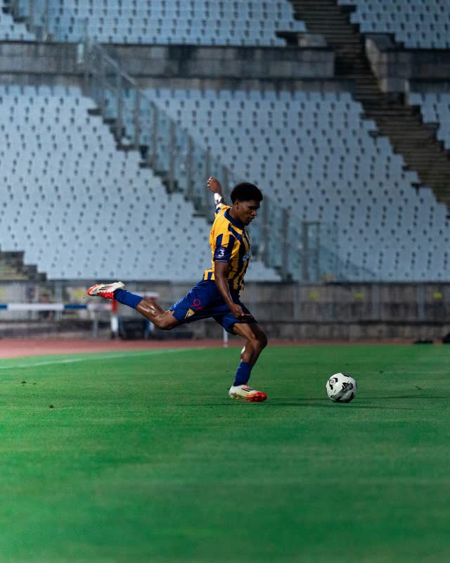 A player in yellow and blue stripes winds up a long pass in front of an empty stand